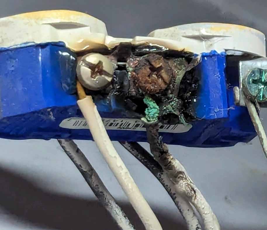 Closeup of electrical outlet removed from wall with wiring still attached showing heat damage and potential fire hazard from arcing activity.